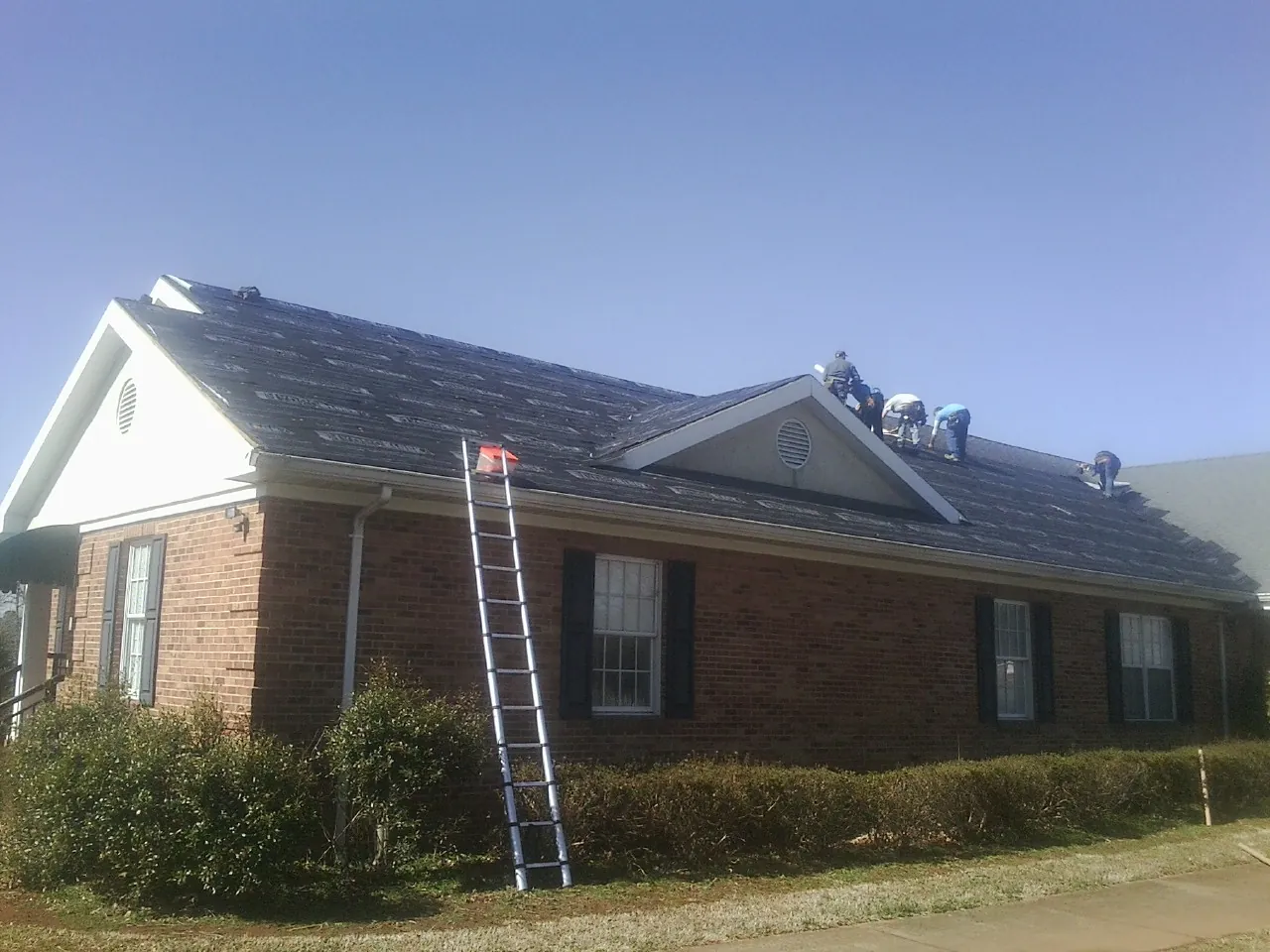 Crew tearing shingles off a brick ranch roof with a ladder set — Emergency Roof Repair work by our crew in Cedar Springs
