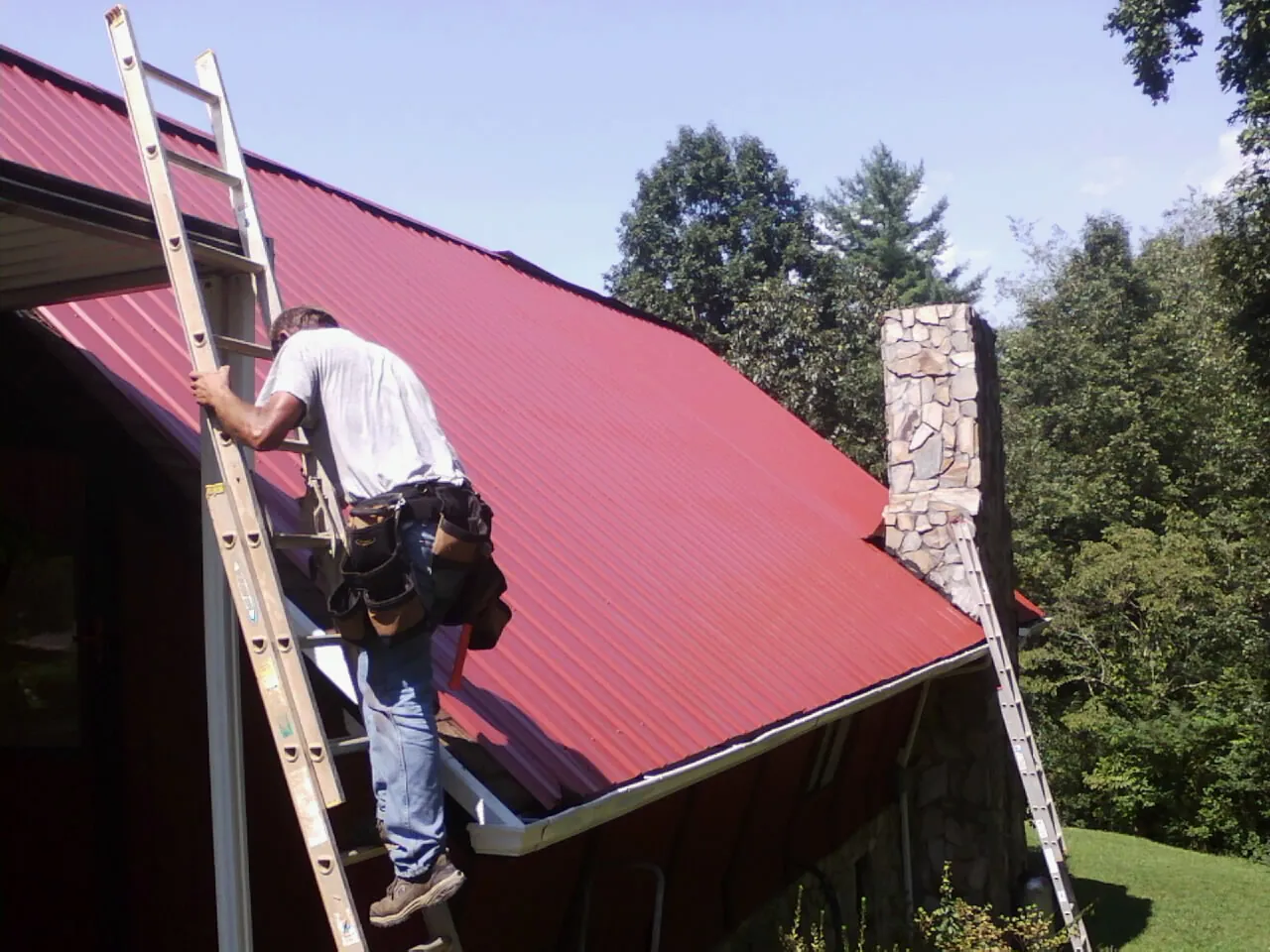 Roofer on a ladder beside a red metal roof and stone chimney — Soffit & Fascia Repair by Roofing Brothers in Cedar Springs